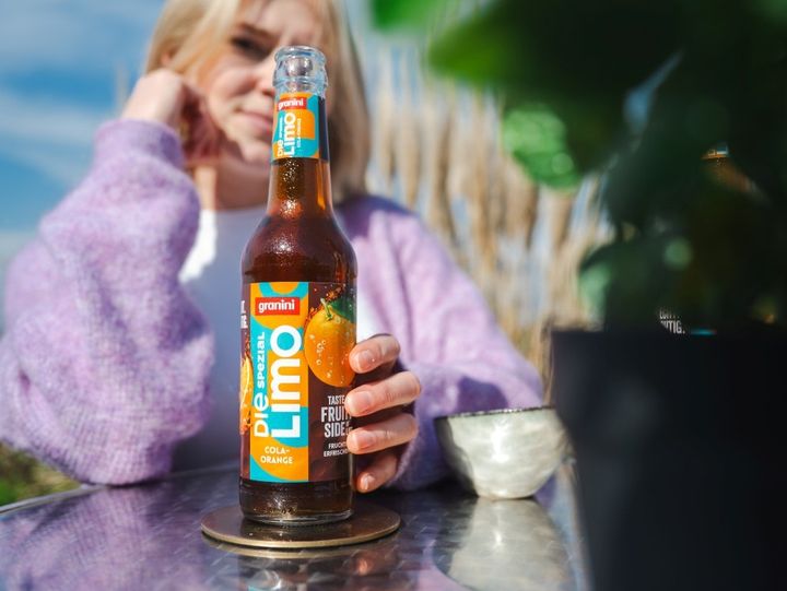 A person wearing a purple sweater holds a bottle of Granini Die Spezial Limo, cola-orange flavor, on a reflective table outdoors.
