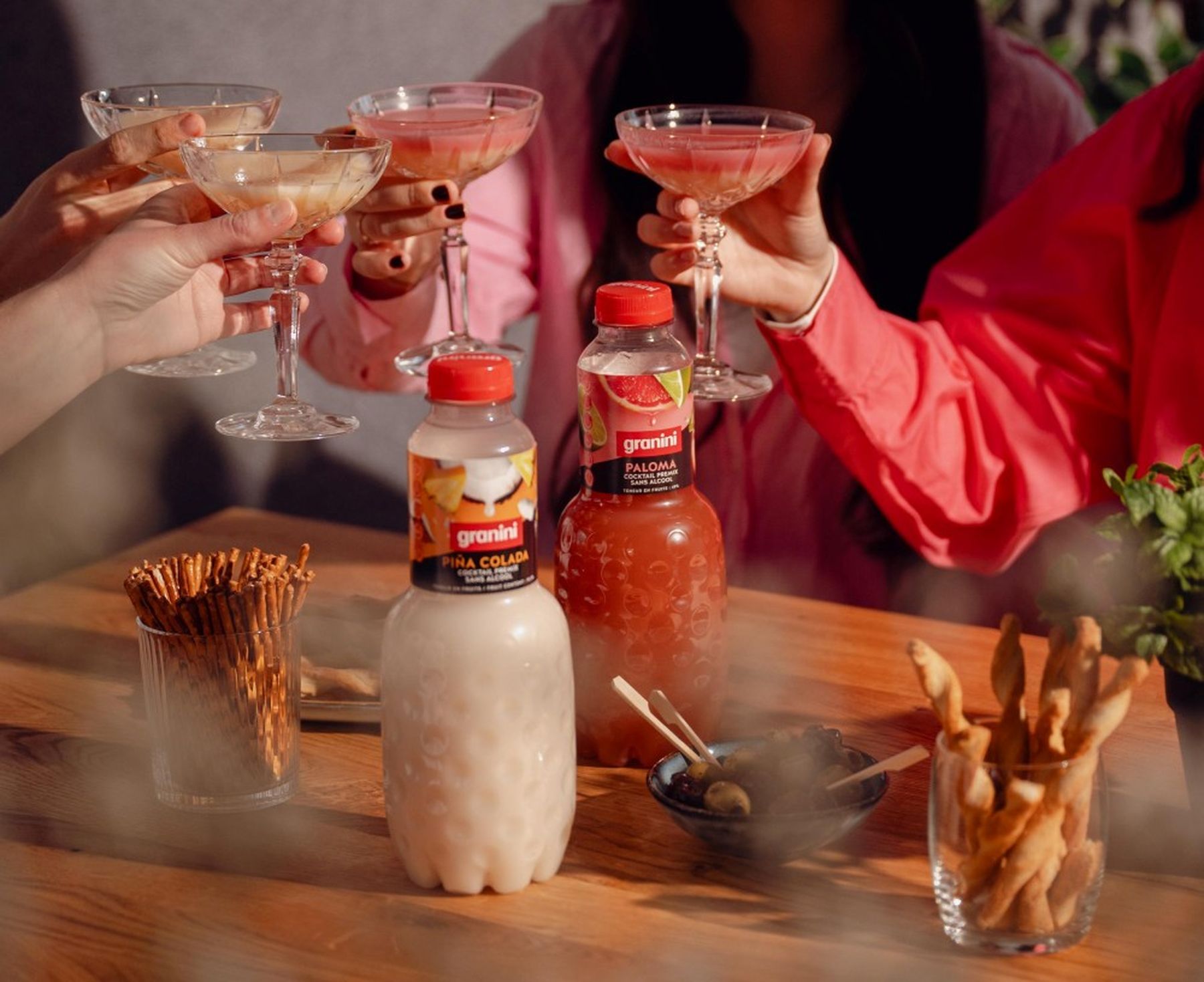 People toasting with drinks in crystal glasses. Bottles of Granini Piña Colada and Paloma sit on a table with snacks.