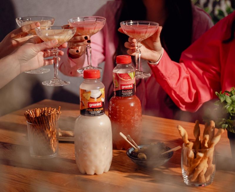 People toasting with drinks in crystal glasses. Bottles of Granini Piña Colada and Paloma sit on a table with snacks.