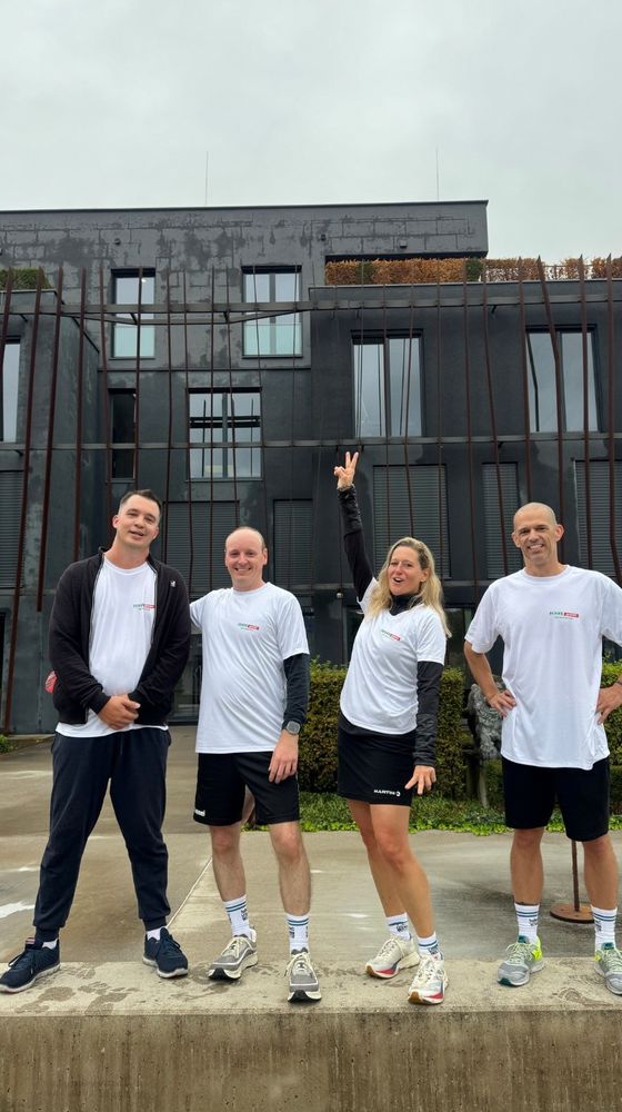 Four people wearing matching white T-shirts and sportswear pose cheerfully in front of a modern building on a cloudy day.