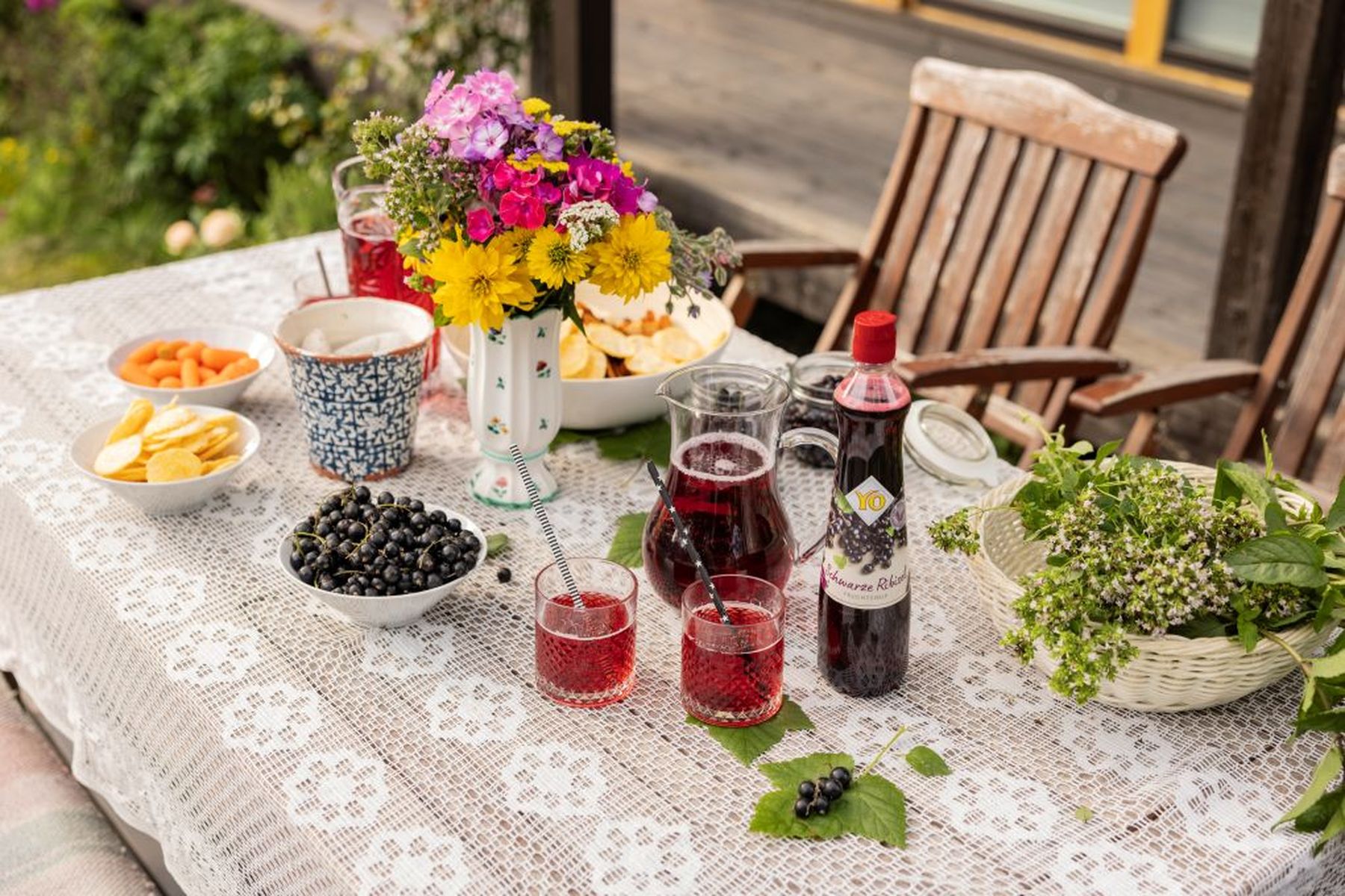 Outdoor table with flowers, bowls of snacks, a bottle, and glasses of red drink on a lace cloth, surrounded by wooden chairs.