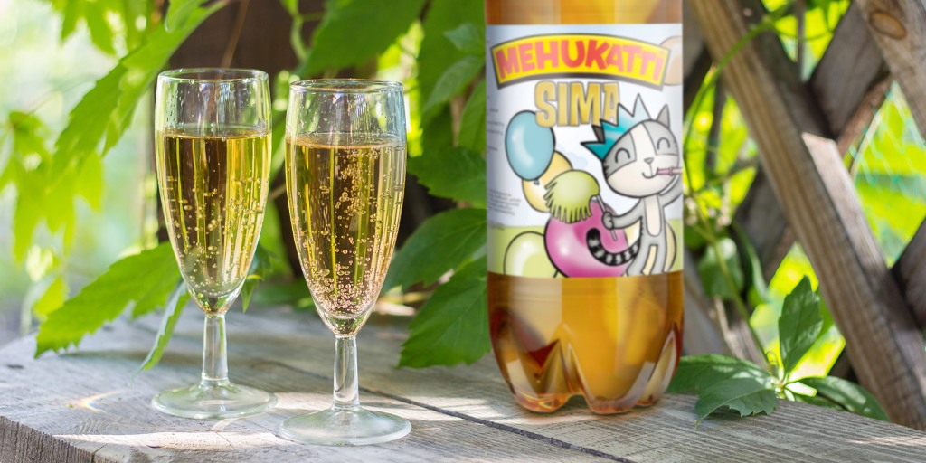 Two champagne flutes filled with bubbly beverage next to a bottle of Mehukatti Sima on a wooden surface, surrounded by green leaves.