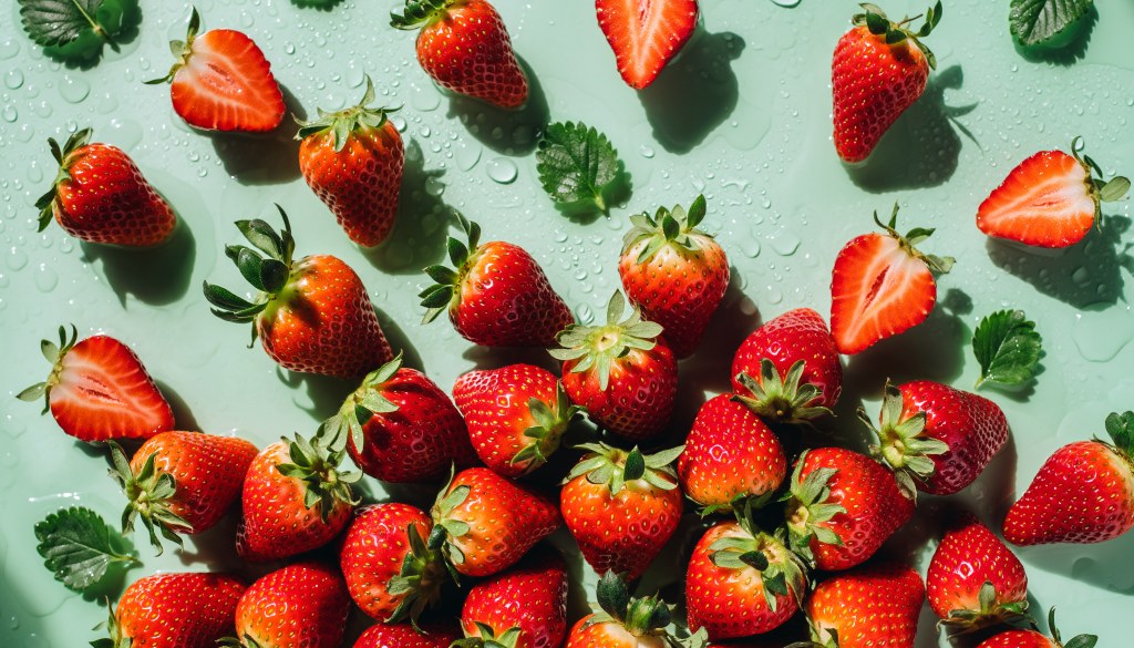 Fresh strawberries, whole and halved, arranged on a light green surface with scattered water droplets and small green leaves.