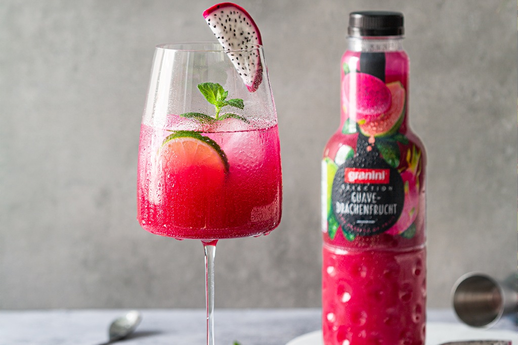 A glass of vibrant pink guava and dragon fruit juice garnished with lime and mint, next to a matching juice bottle on a gray background.