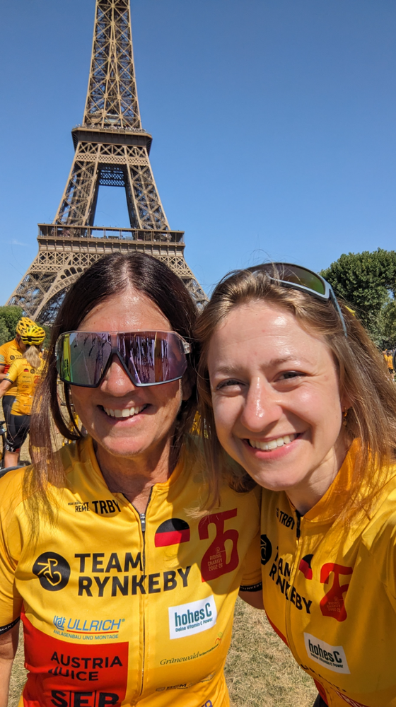 Two women in yellow cycling jerseys smiling in front of the Eiffel Tower with other cyclists in the background.