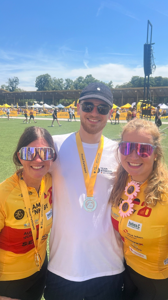 Three people wearing sunglasses and medals smile on a sunny athletic field, surrounded by event tents and participants in yellow shirts.