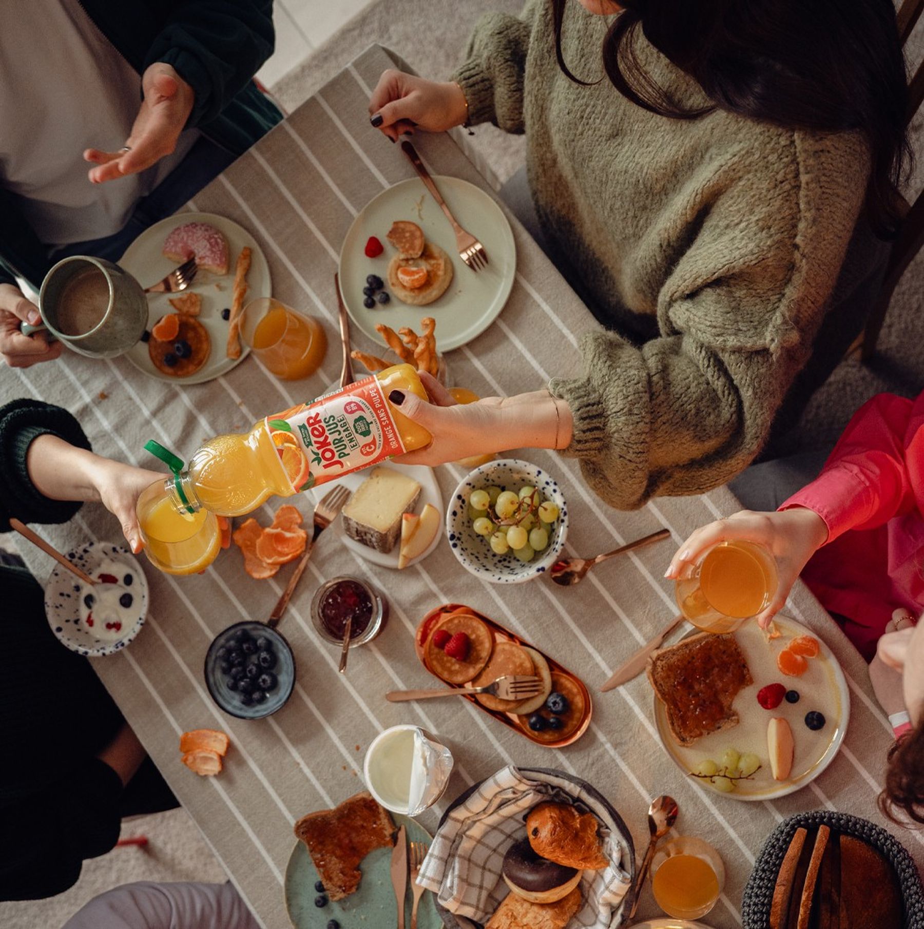 Top view of friends having breakfast with pancakes, fruit, yogurt, and juice on a striped tablecloth. One person pours orange juice into a glass.