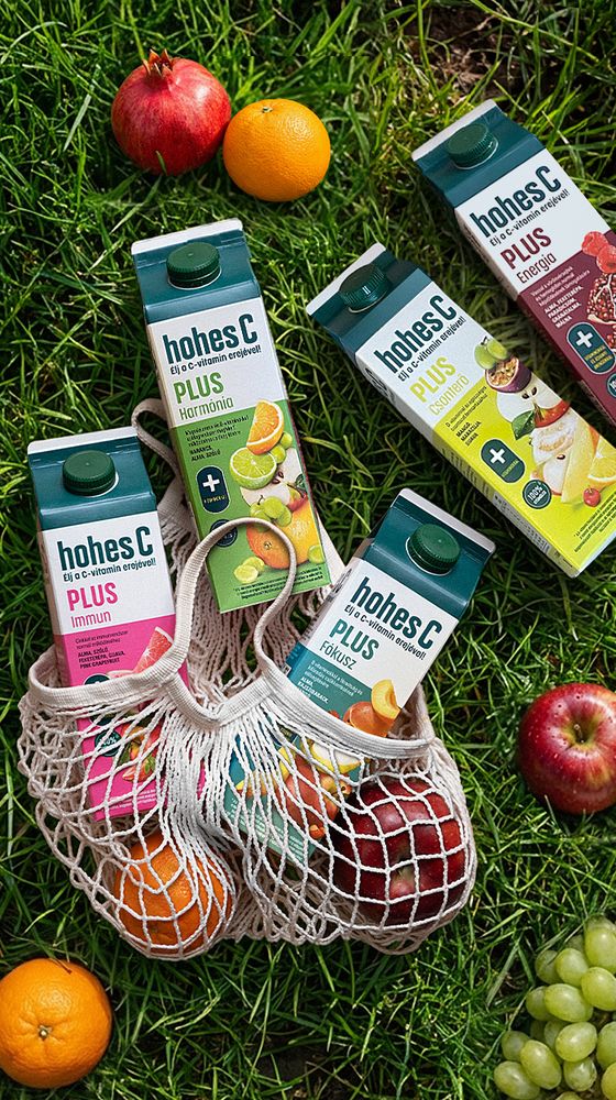 Several cartons of Hohes C juice and fresh fruits, including apples and oranges, lie on grass with some in a netted bag.
