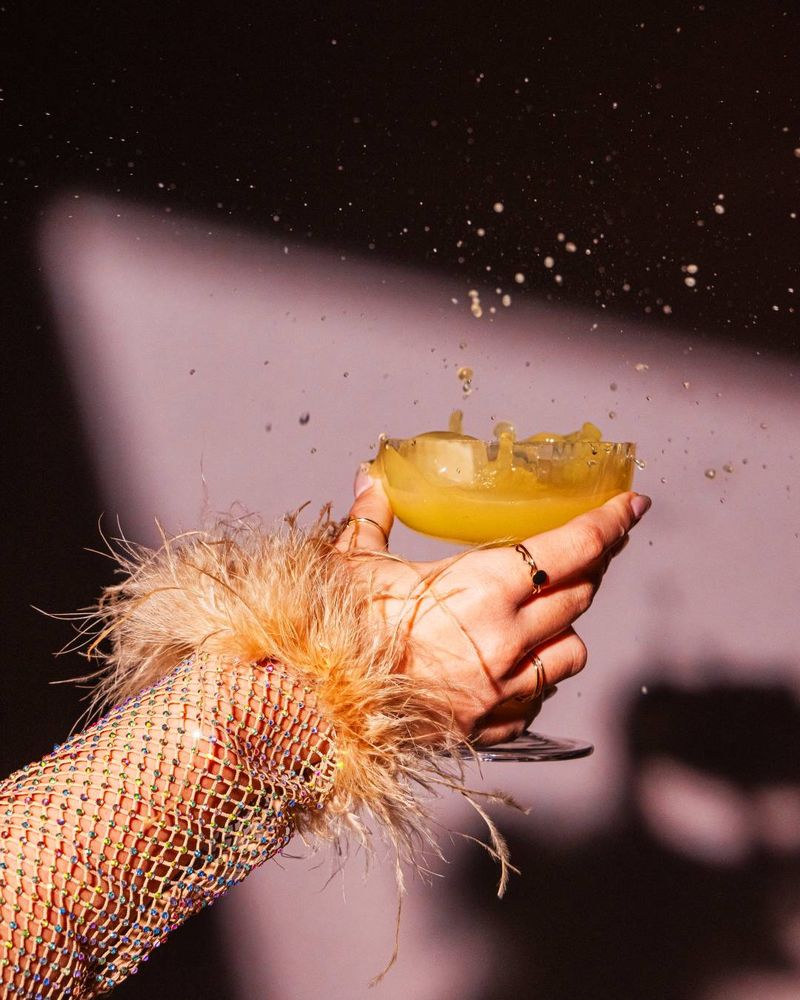 A hand holding a cocktail glass, spilling yellow liquid. The person wears a sequined, feathered sleeve. Shadows cast on a pink wall.