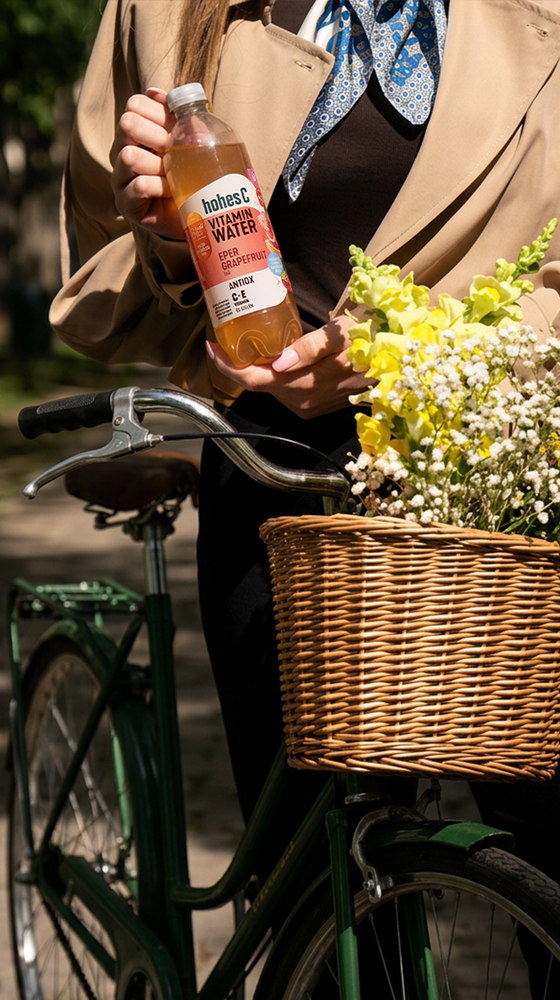 Person holding a bottle of vitamin water stands by a green bicycle with a wicker basket filled with yellow and white flowers.