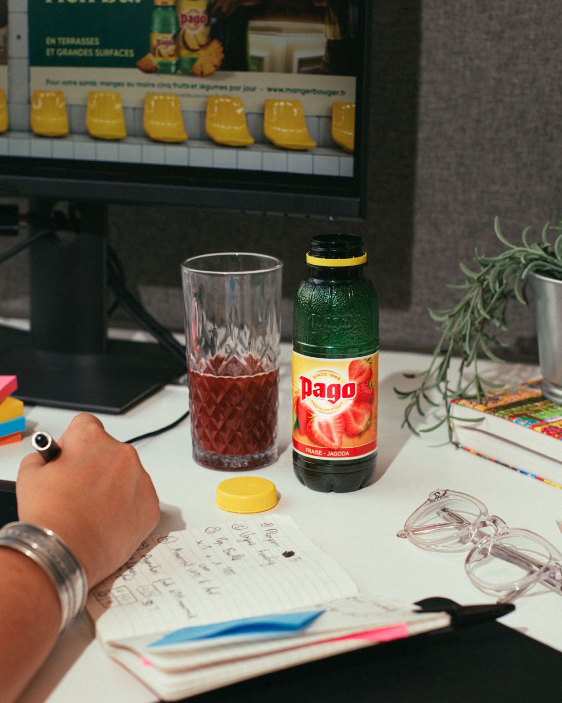A person writes in a notebook at a desk with a Pago juice bottle, glass of juice, computer monitor, and stack of books.