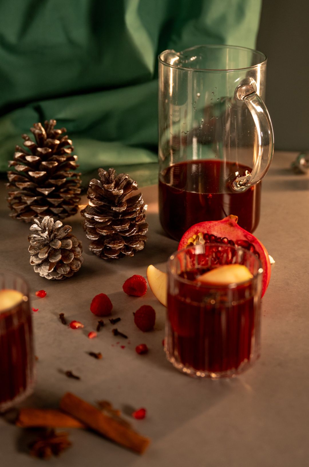 Pitcher and glass of spiced red mulled wine with apple slice, pomegranate, raspberries, cloves, cinnamon sticks and pinecones.