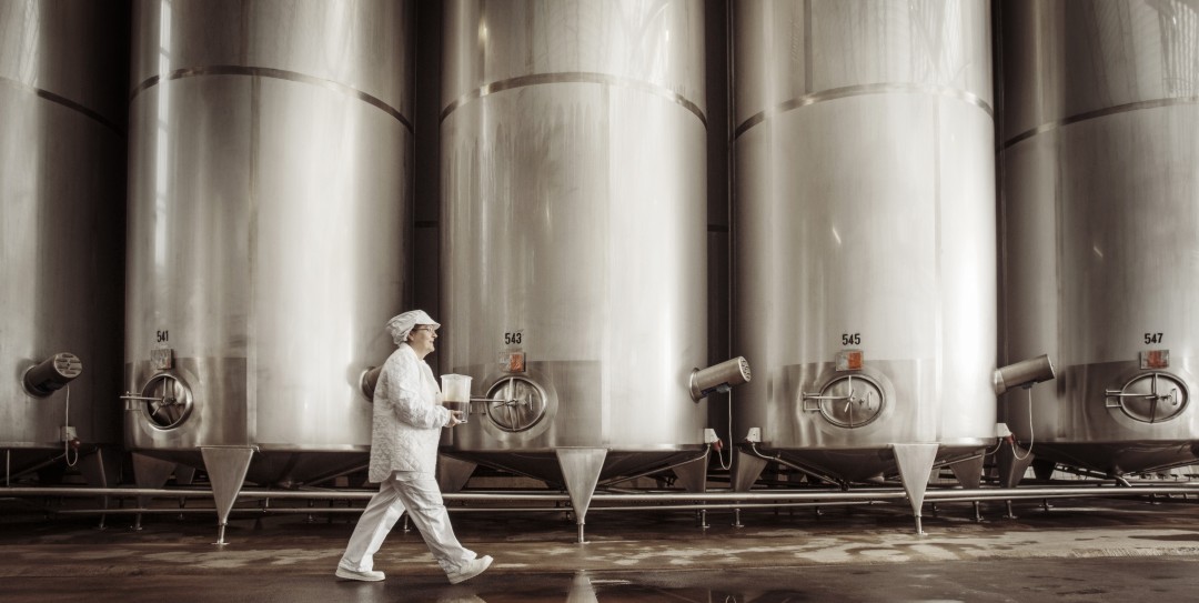 Person in white uniform walking past large stainless steel tanks in an industrial setting, possibly a brewery or dairy facility.