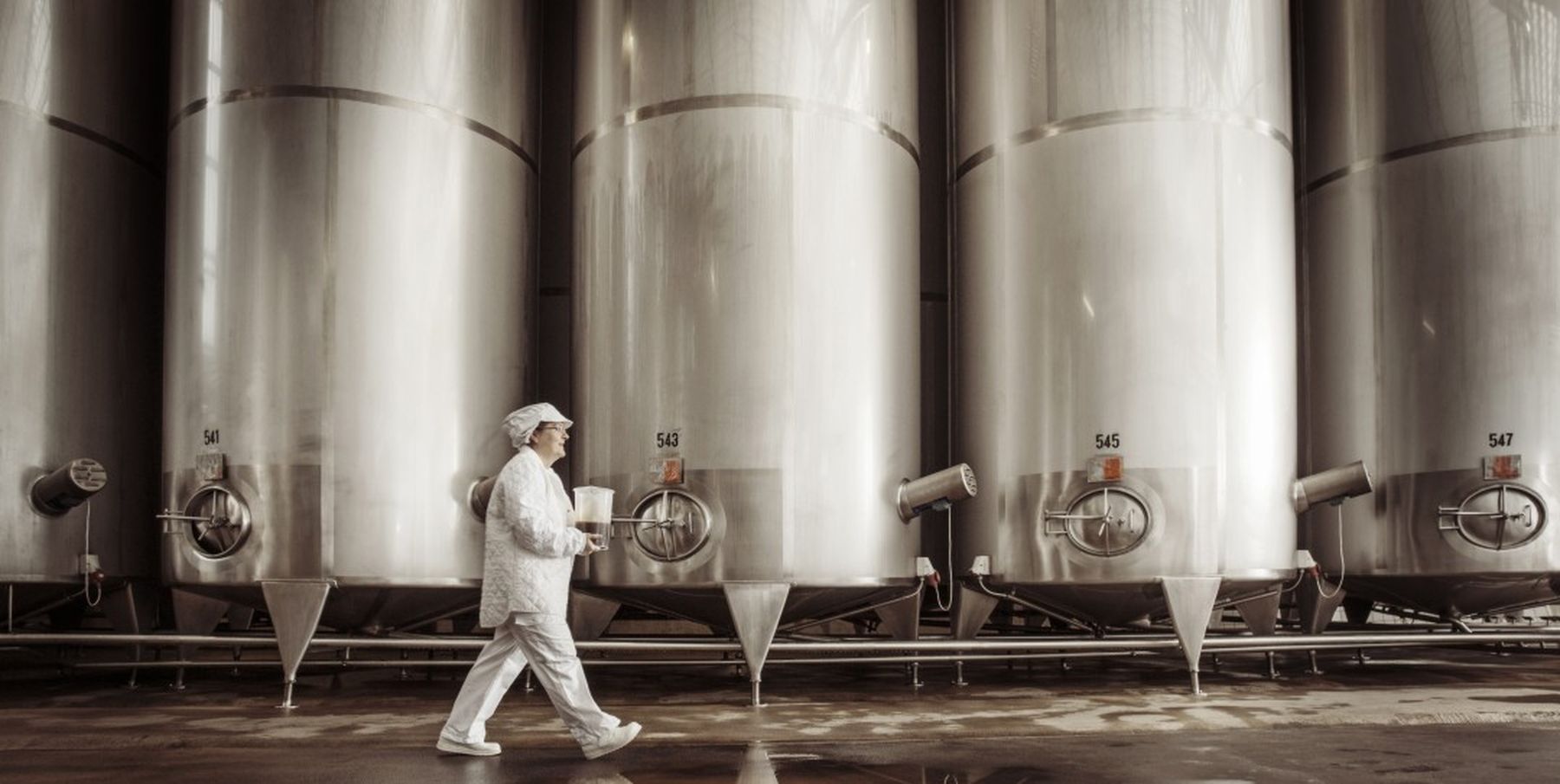 Person in white uniform walking past large stainless steel tanks in an industrial setting, possibly a brewery or dairy facility.