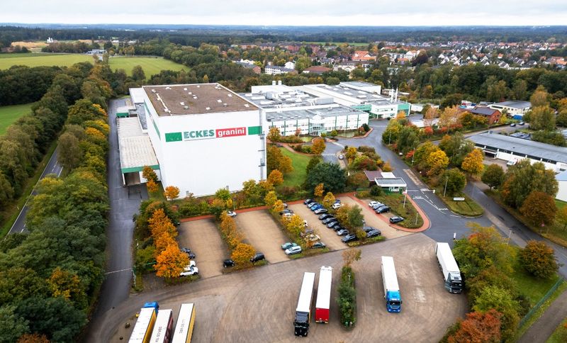 Factory Bad Fallingbostel: Aerial view of a large industrial complex surrounded by trees with autumn foliage, featuring several parked trucks and nearby buildings.