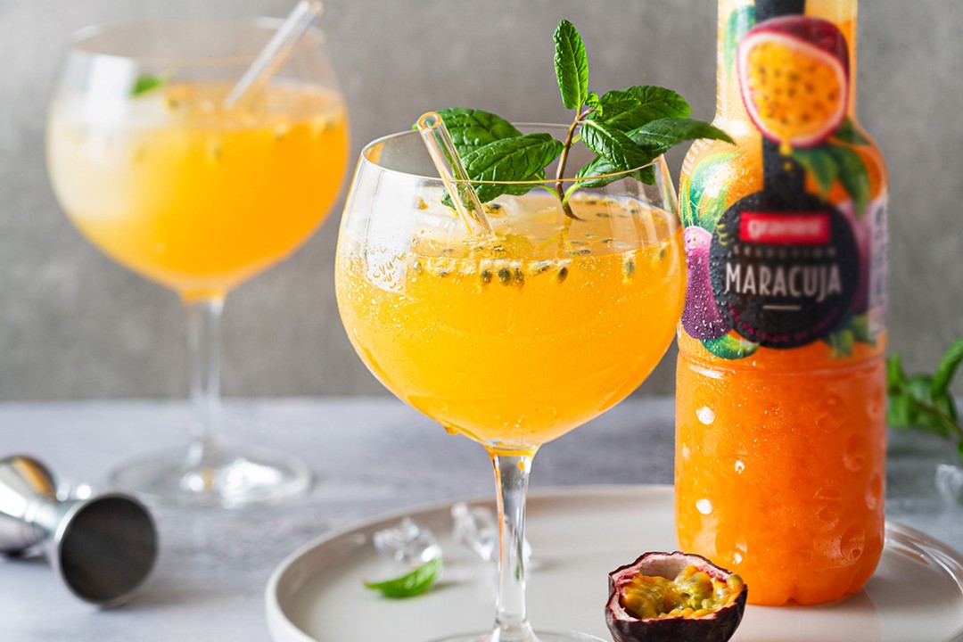 Two glasses of vibrant orange passion fruit juice garnished with mint, alongside a bottle of "Maracuja" juice and a halved passion fruit.