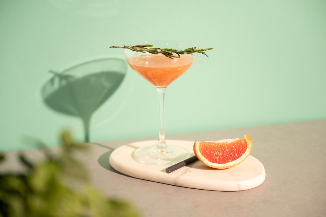 Pink grapefruit cocktail in a coupe glass garnished with rosemary, grapefruit wedge on a marble coaster.