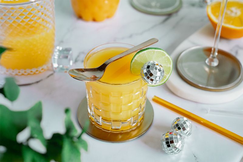 Margarita with orange juice and a lime slice and spoon on top, surrounded by disco ball decorations on a marble surface.