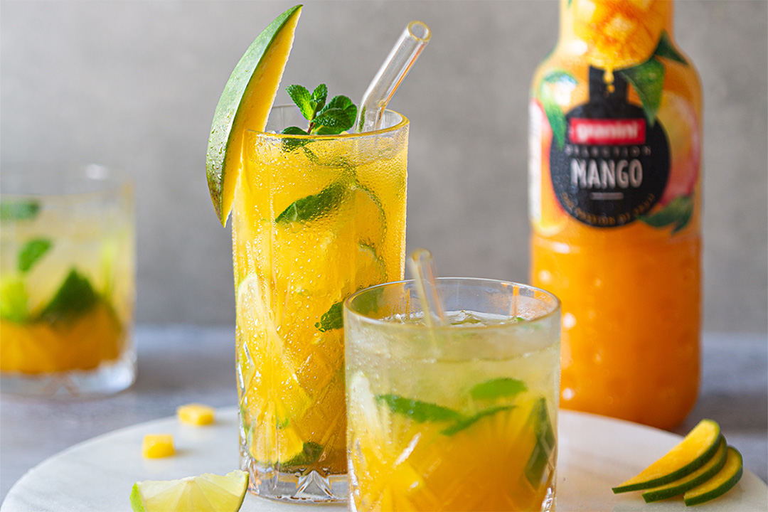 Two glasses of mango mojito garnished with mint and lime slices, with a bottle of granini mango juice in the background.