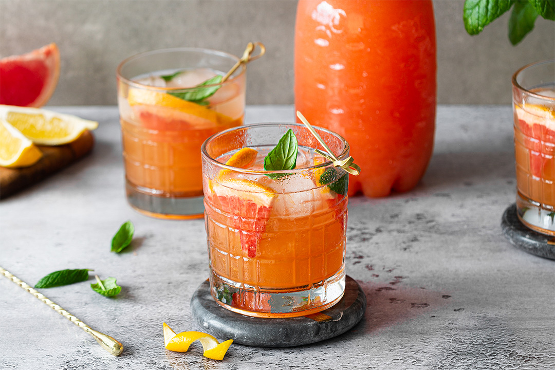 Two glasses of iced grapefruit cocktail with mint and citrus slices, a bottle of juice, and garnish on a gray surface.