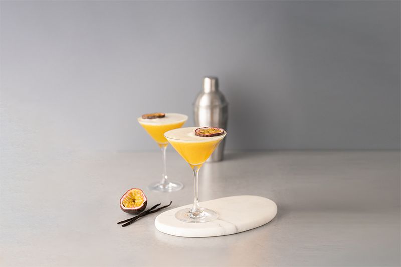 Two passion fruit cocktails in martini glasses on a white board, with a cocktail shaker and sliced passion fruit nearby.