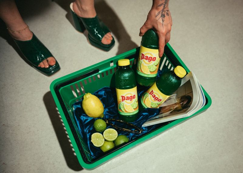 A person with green shoes places a bottle of Pago citrus drink in a basket containing limes, a lemon, vanilla beans, and a magazine.
