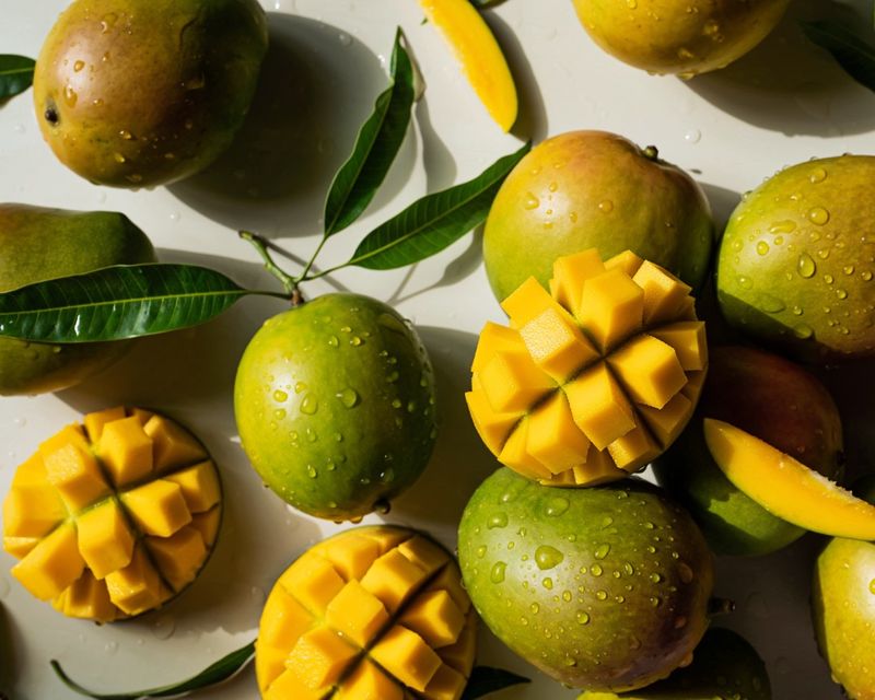 Mangos with green skins and water droplets, surrounded by sliced mango cubes and green leaves on a white surface.
