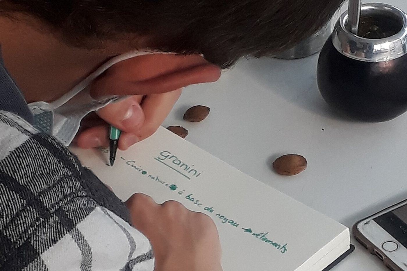 Person writing "granini" in a notebook with a pen, next to a gourd with a metal straw and scattered nuts on a table.
