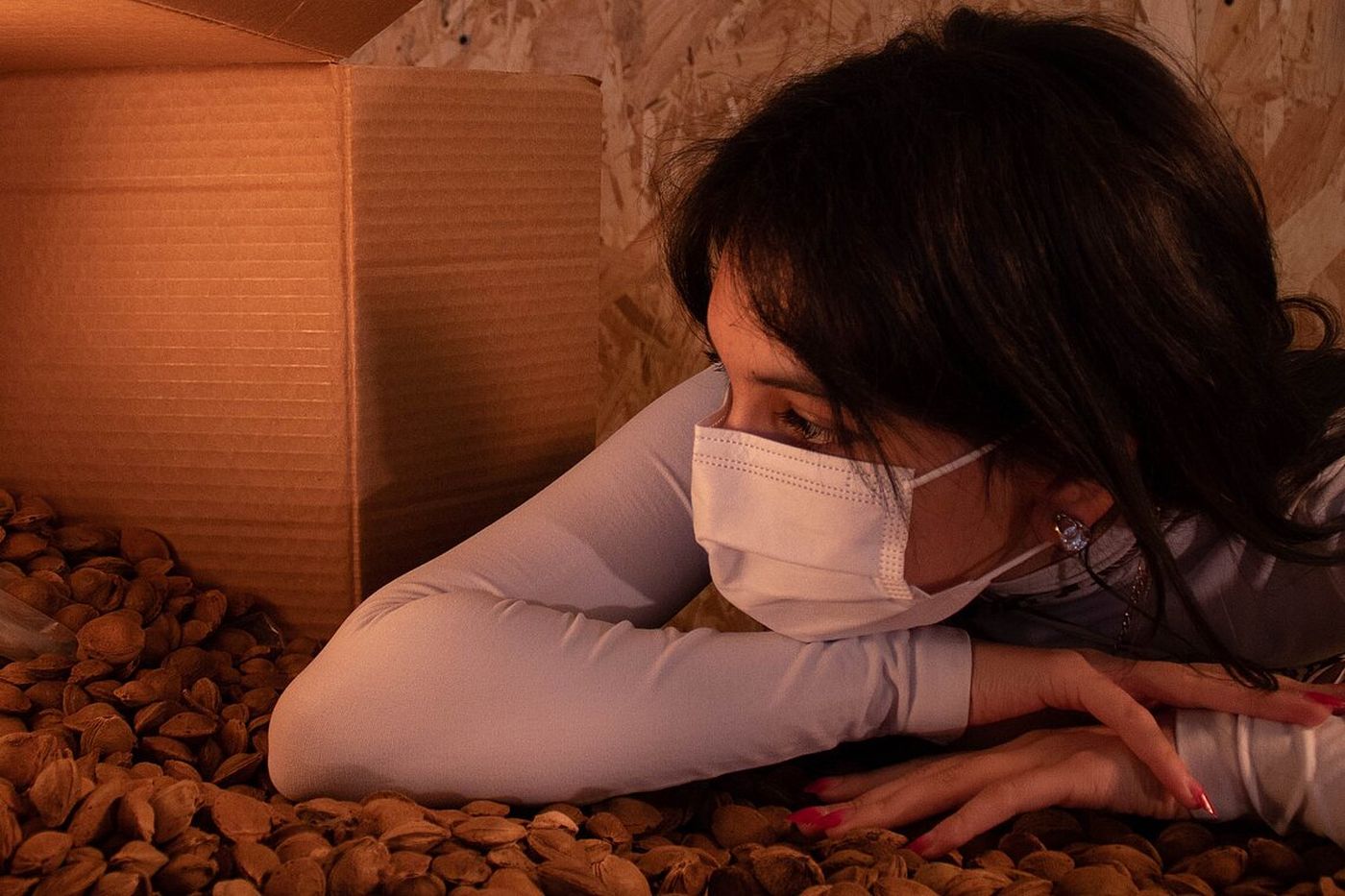 Woman in a mask leans on a pile of almonds next to an open cardboard box, looking thoughtfully to the side.