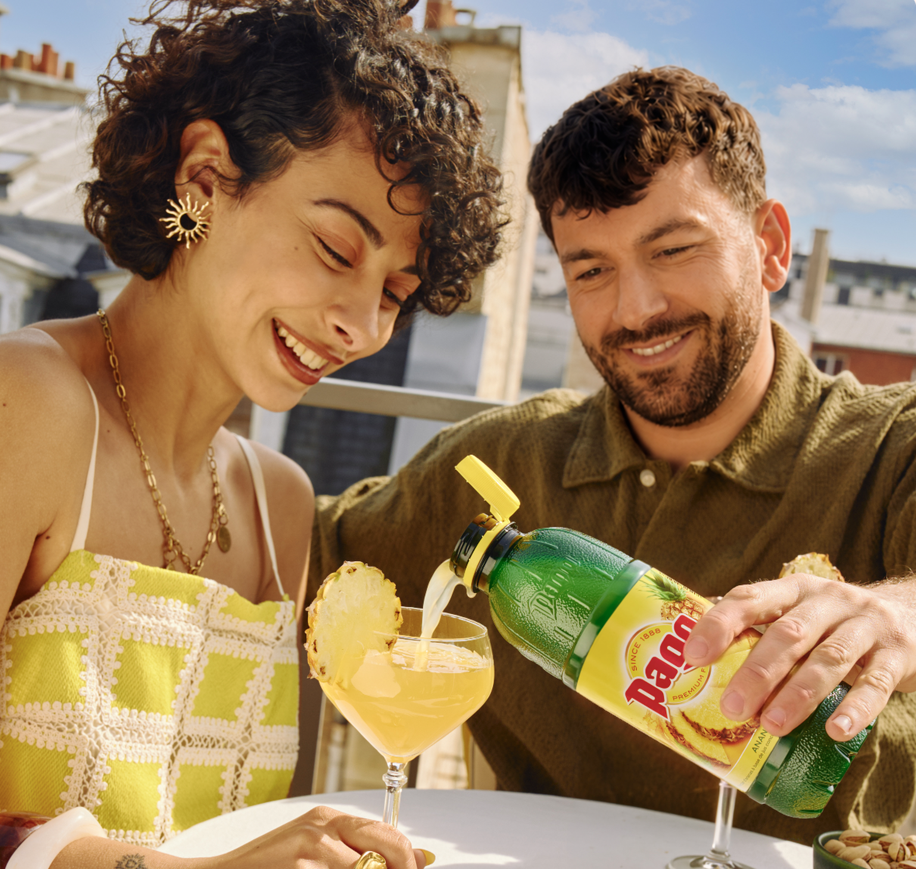 Un couple souriant en date sur un rooftop parisien. l'homme sert un Pago Ananas 75cl à son date