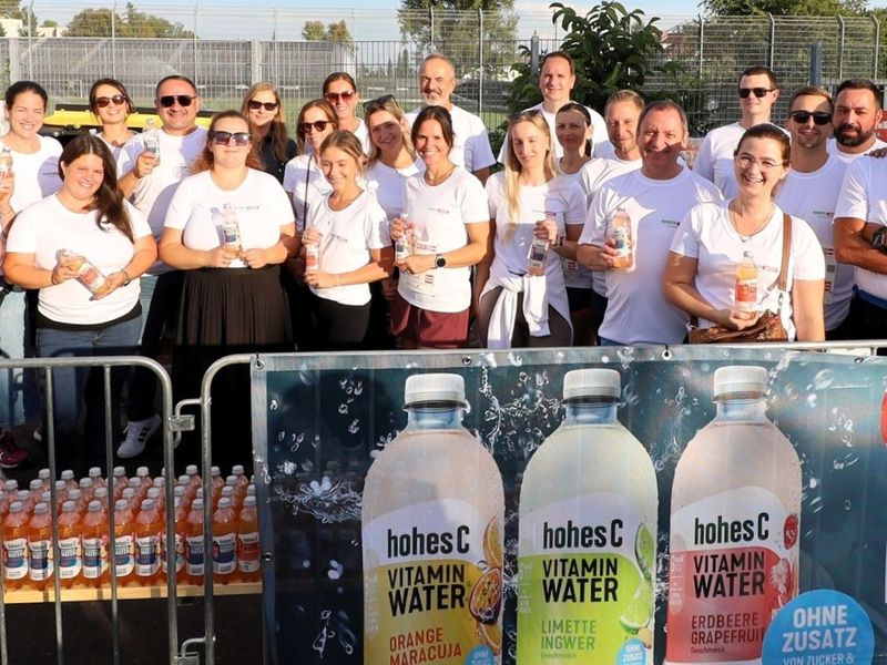 A group of people in white shirts holding bottles of vitamin water, standing behind a banner displaying different flavors.