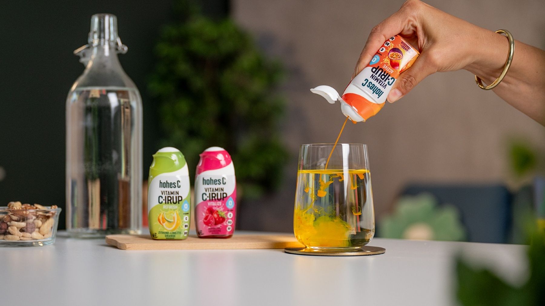 A person pours hohes C Vitamin C-IRUP into a glass of water. Green and red syrup bottles and a glass water bottle are in the background.