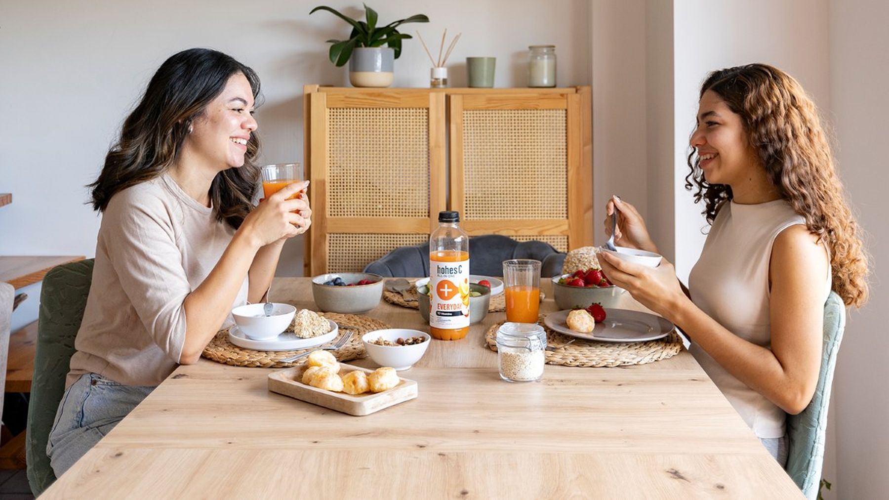 Two women enjoying breakfast at a table with cereal, pastries, fruit, and hohes C ALL-IN-ONE. A plant is in the background.