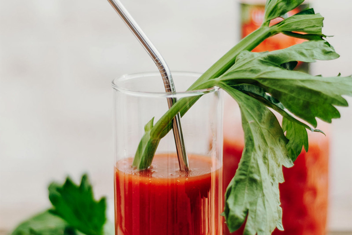 Bloody Mary with a metal straw and celery stalk garnish, set against a blurred background with a bottle partially visible.