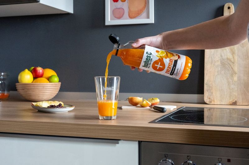 Person pouring orange juice from a bottle into a glass, with fruit on the counter and a cutting board in the background.