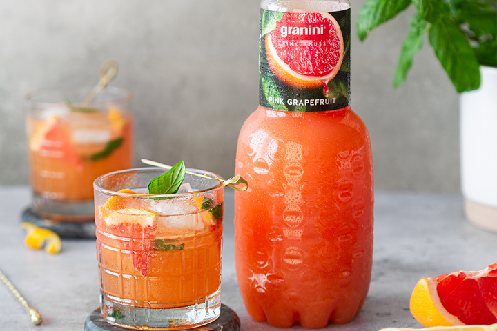 Bottle of pink grapefruit juice beside a glass with a sliced garnish and mint leaves on a table.