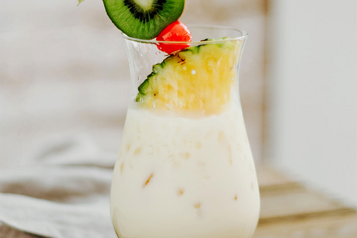 A creamy cocktail garnished with a slice of pineapple, cherry, and kiwi on a wooden table, set against a softly blurred background.