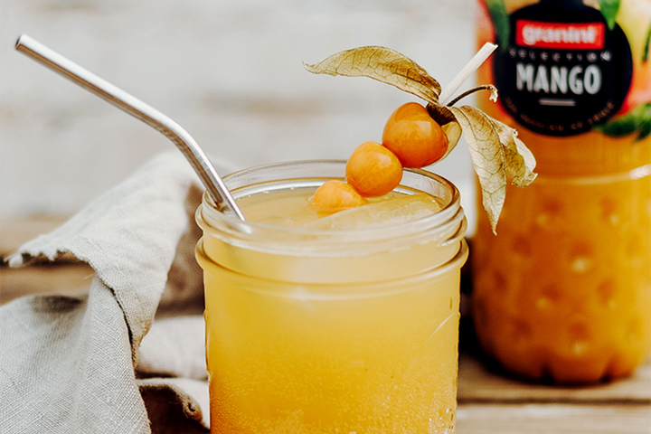 A mason jar with mango juice, ice, a metal straw, and garnished with physalis. A mango juice bottle is in the background.