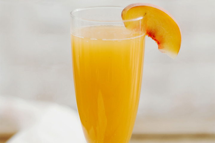 A glass of bellini with orange juice garnished with a fresh peach slice on the rim.