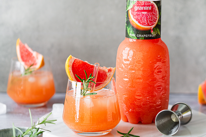 Bottle of granini pink grapefruit juice beside a glass with ice, grapefruit slice, and rosemary sprig, with a jigger on the table.