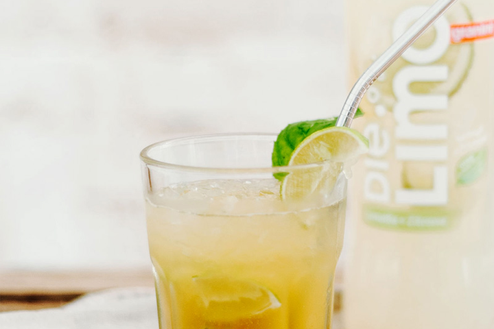 A glass of iced lemonade with a lime slice on the rim and a metal straw, against a light background with a lemonade bottle partially visible.