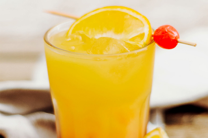 A glass of tequila sunrise, garnished with an orange slice and a cherry on a skewer, placed on a light-colored cloth.