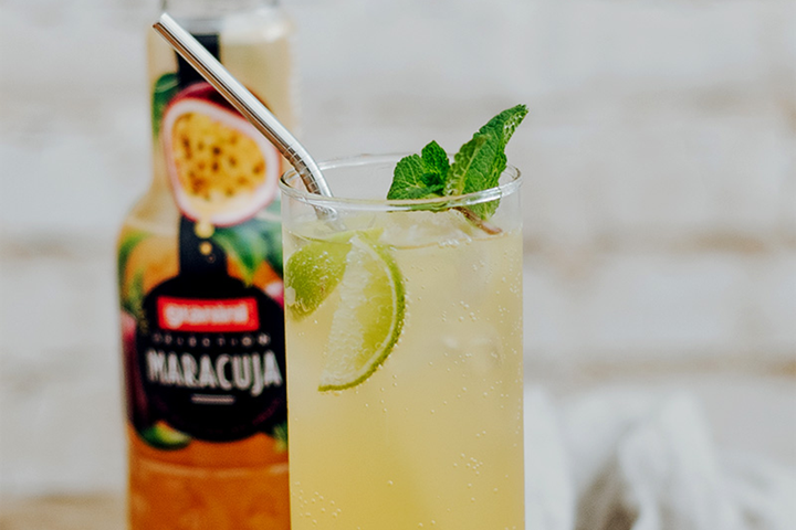 Chilled passion-fruit cocktail in tall glass with ice, lime slices, mint and metal straw; blurred maracuja bottle in background.