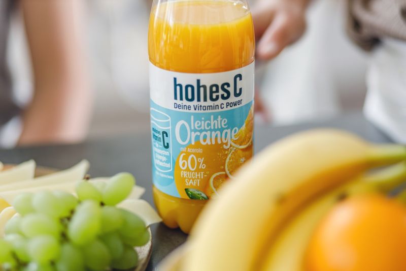 A bottle of hohes C orange juice on a table with bananas and green grapes in the foreground.
