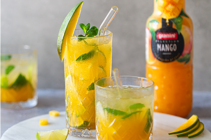 Two refreshing mango mint cocktails with lime wedges, one garnished with a mango slice, beside a bottle of mango juice on a light-colored surface.