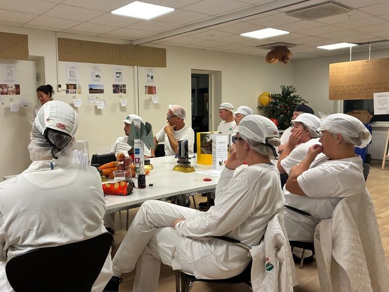 A group of people in white uniforms and hairnets attentively listening to a presentation in a room with informational posters on the walls.