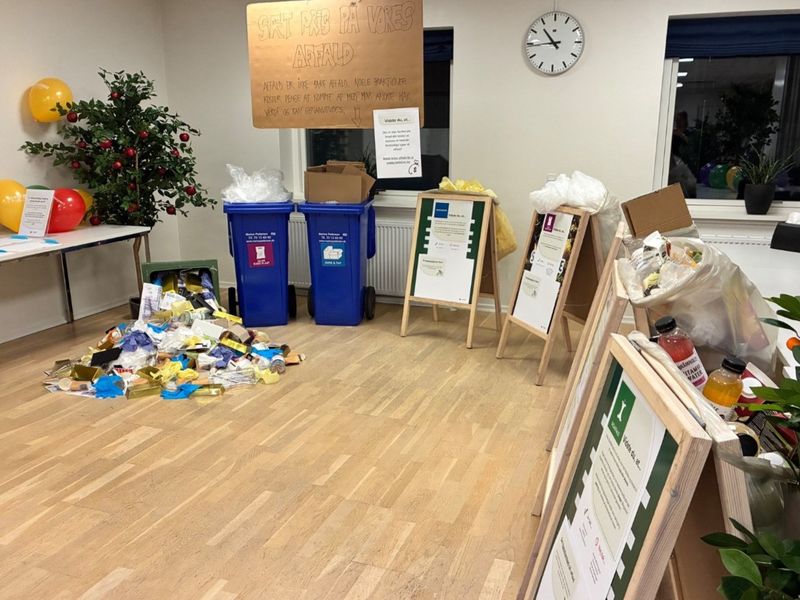 Room setup with recycling bins, a pile of sorted waste, informational posters on easels, colorful balloons, and a clock on the wall.