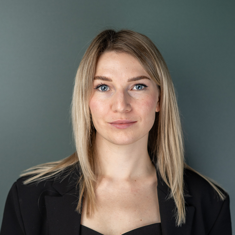 A woman with long blonde hair wearing a black blazer, standing against a neutral background, looking directly at the camera with a calm expression.