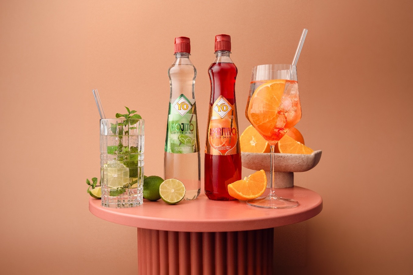 Two bottles YO Cocktails labeled "Mojito" and "Aperitivo" on a table with cocktails, lime, mint, and orange slices against an orange background.