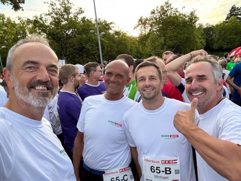 Four men in white shirts, smiling and posing for a selfie at an outdoor event. One man gives a thumbs-up. Trees are visible in the background.